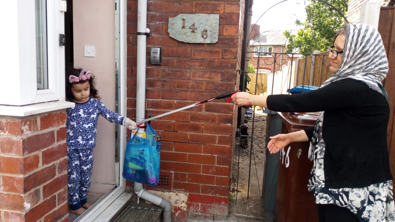 A woman hands a plastic bag of jigsaws and crafts - using a litter picker - to a child stood in a doorway, wearing plastic gloves