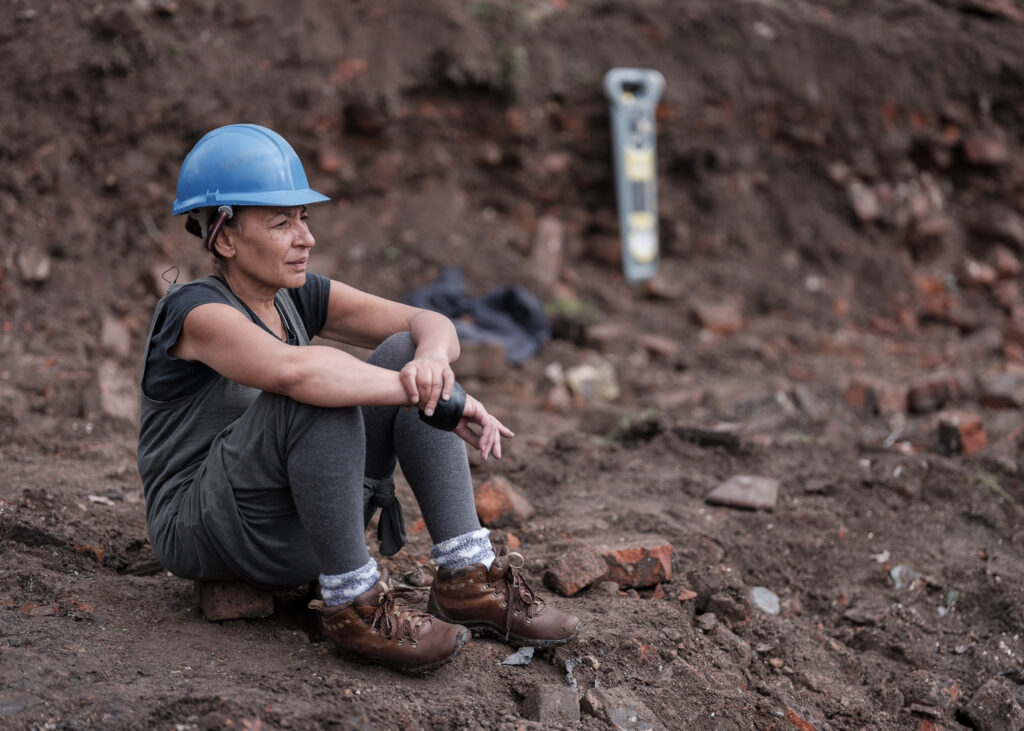 Linda Brogan in a builder's hat sat on a brick at excavation of the Reno surrounded by rubble