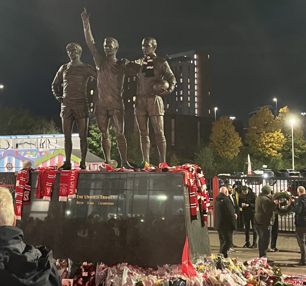 A statue of Sir Bobby Charlton, Denis Law and George Best with scarves adorning it.