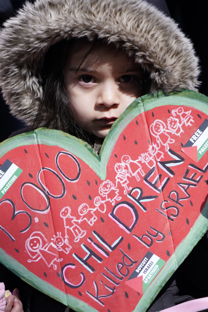 A child holds a heart shaped poster reading 13,000 children killed by Israel, calling for a ceasefire.