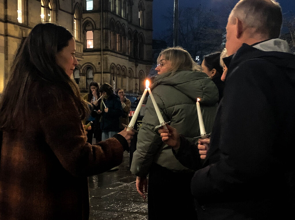 A young woman holding a candle uses it to light the candle of an elderly couple, smiling. The two candles are joined together in the centre of the image. There is a small crowd of attendees in the background, and everyone is wearing coats. It is dark outside and the pavement is wet with rain.