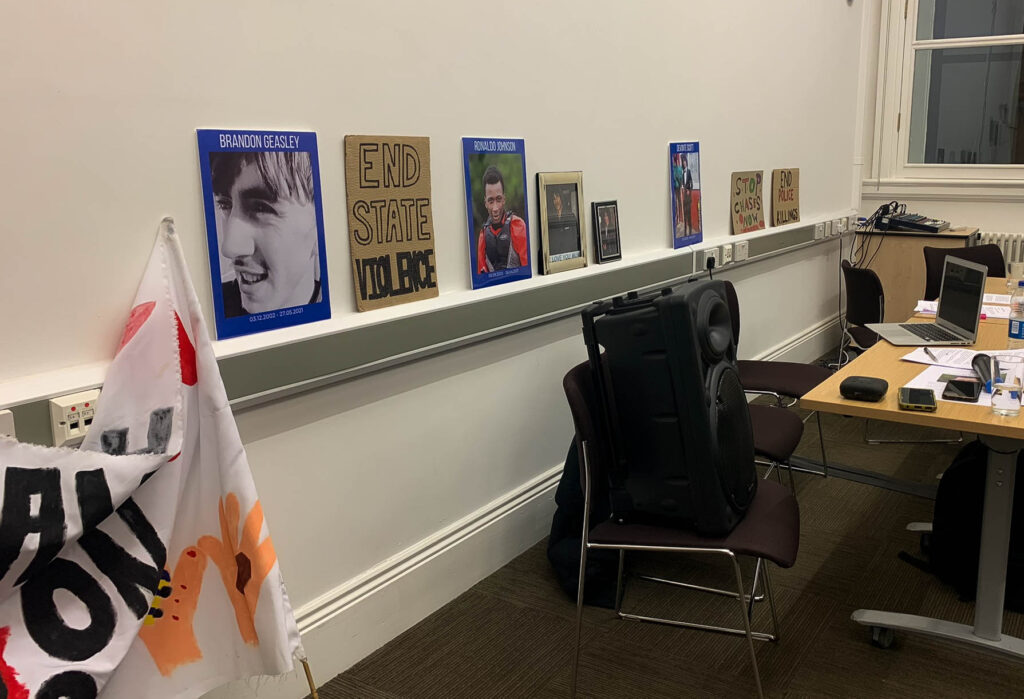 An office in Friends Meeting House, featuring a desk, chairs, a laptop, and a speaker. On the wall is a line of cardboard signs and family photos of Brandon Geasley, Ronaldo Johnson, and Devonte Scott. There is a folded banner in the foreground, which unreadable. The cardboard signs say: "End state violence", "Stop chases now", and "End police killings".