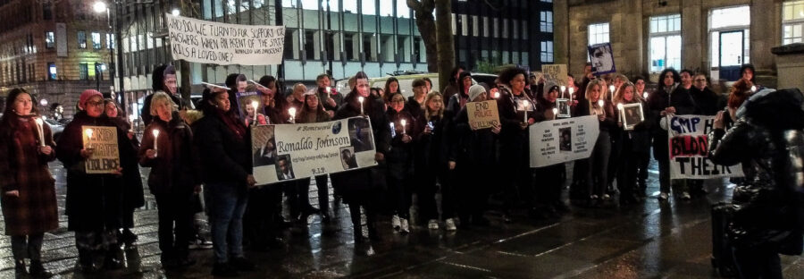 Families and attendees stand in a crowd outside Friends Meeting House, holding banners, lit candles, and homemade carboard signs. They are all wearing coats. The sky is dark, and the pavement is wet from rain. The visible signs say: "End state violence", "End police killings", and "End police pursuits". The visible banners say: "Who do we turn to for support or answers when an egent of the state kills a loved one? Ronaldo was innocent", "#RonsWorld. Ronaldo Johnson. R.I.P.", and "G.M.P. have blood on their hands."