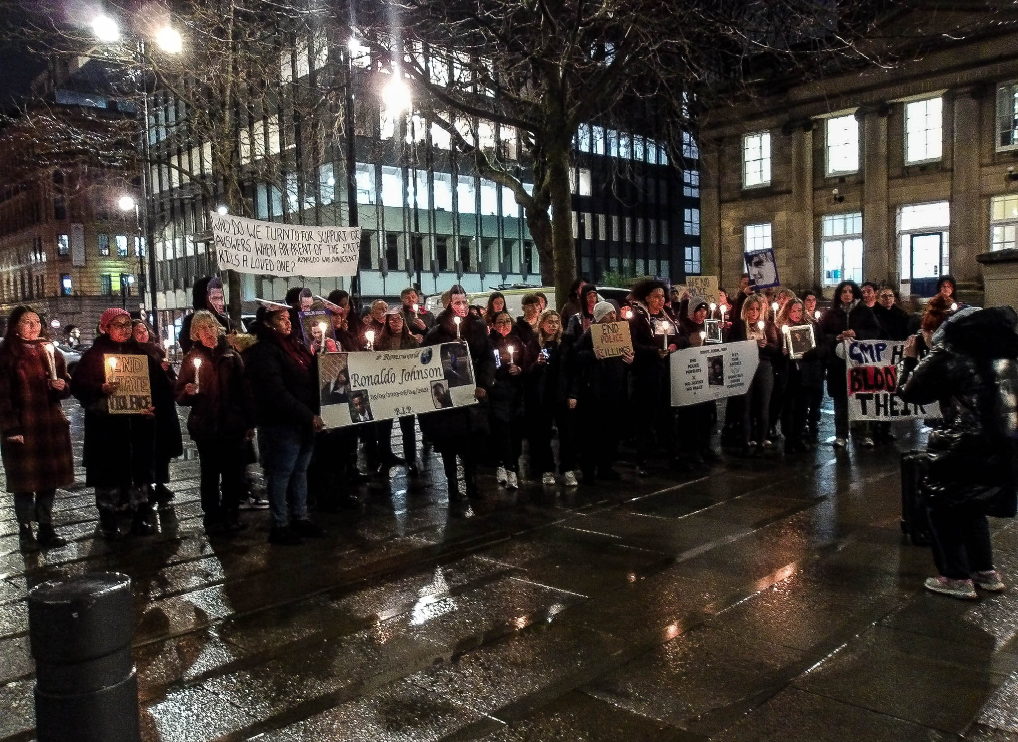 Families and attendees stand in a crowd outside Friends Meeting House, holding banners, lit candles, and homemade carboard signs. They are all wearing coats. The sky is dark, and the pavement is wet from rain. The visible signs say: "End state violence", "End police killings", and "End police pursuits". The visible banners say: "Who do we turn to for support or answers when an egent of the state kills a loved one? Ronaldo was innocent", "#RonsWorld. Ronaldo Johnson. R.I.P.", and "G.M.P. have blood on their hands."