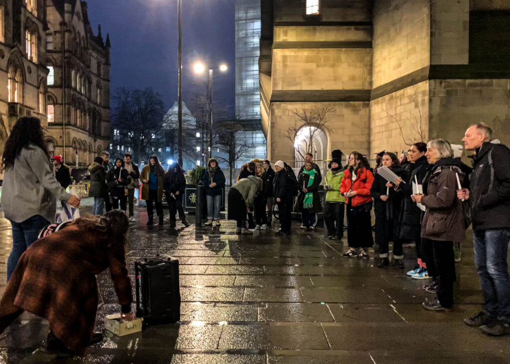 A large crowd formed outside Friends Meeting House, all wearing coats. Zara Manoehoetoe has her back to the camera and is addressing the crowd with a microphone. The sky is dark, and the pavement is wet with rain. The crowd is listening attentively. 