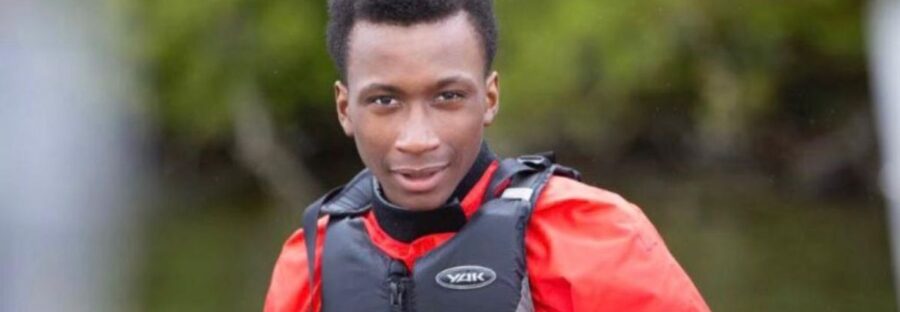 Headshot of Ronaldo Johnson. He is a Black teenage male wearing a red waterproof jacket and black life jacket, staring directly into the camera and smiling. Behind him is a river and green foliage. He is in a canoe, but the canoe has been cropped out.