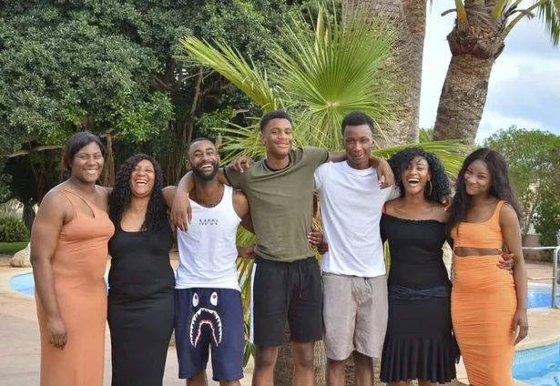 Ronaldo Johnson with his his mother, three sisters, and two brothers, all smiling for the camera with their arms around each other. He is third from the right, wearing a white t-shirt and beige shorts. He is looking directly into the camera and smiling with his arms around his brother and sister, who are hugging him back. In the background is a swimming pool, palm trees, and a blue sky. 