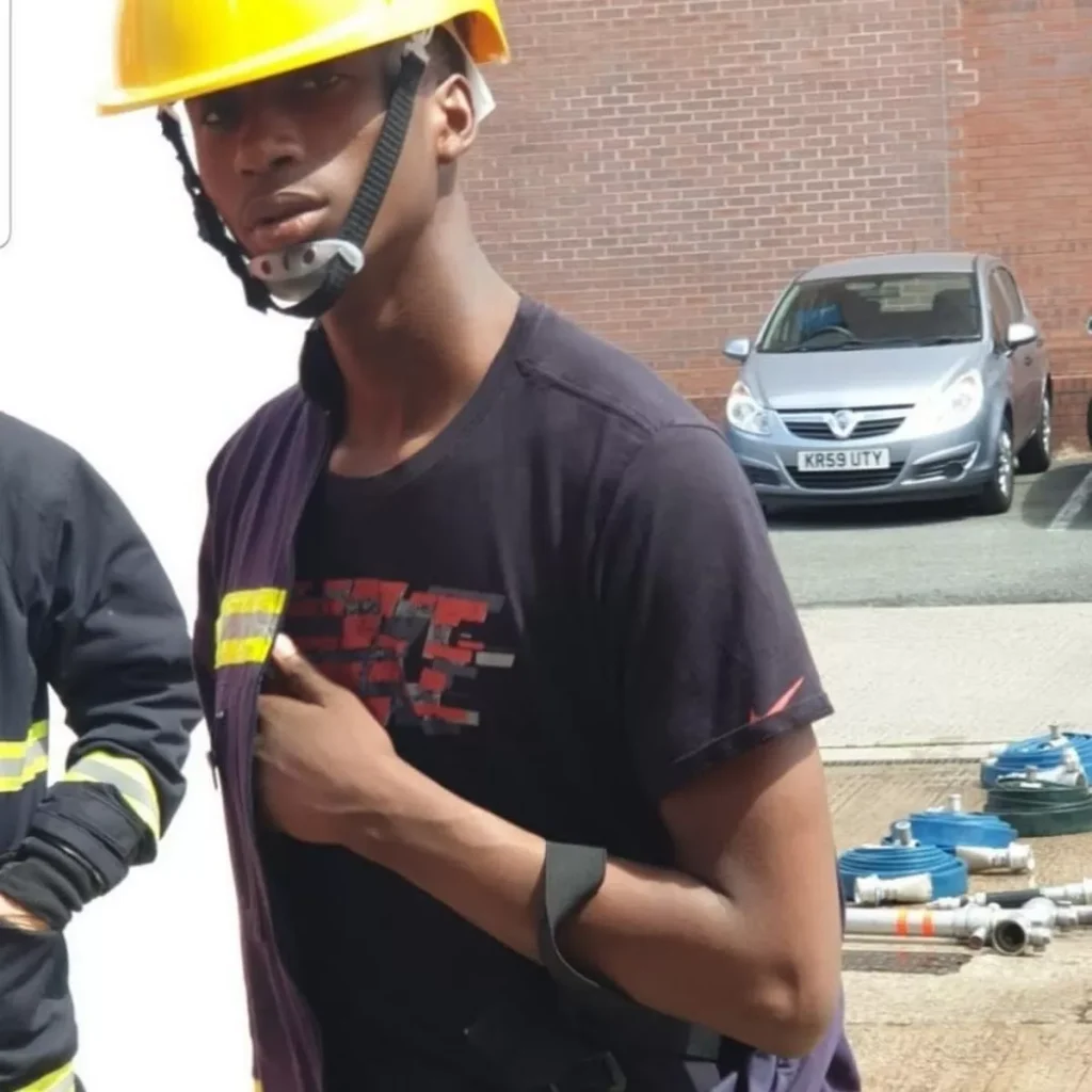 Candid photo of Ronaldo Johnson wearing a construction uniform. He wears a yellow hard hat and a dark T-shirt, and appears to be halfway taking off a dark jacket with a high-vis strip. He is looking directly into the camera. The weather is sunny and there are some disassembled scaffolding pipes on the ground behind him.