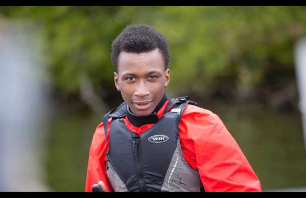 Headshot of Ronaldo Johnson. He is a Black teenage male wearing a red waterproof jacket and black life jacket, staring directly into the camera and smiling. Behind him is a river and green foliage. He is in a canoe, but the canoe has been cropped out.