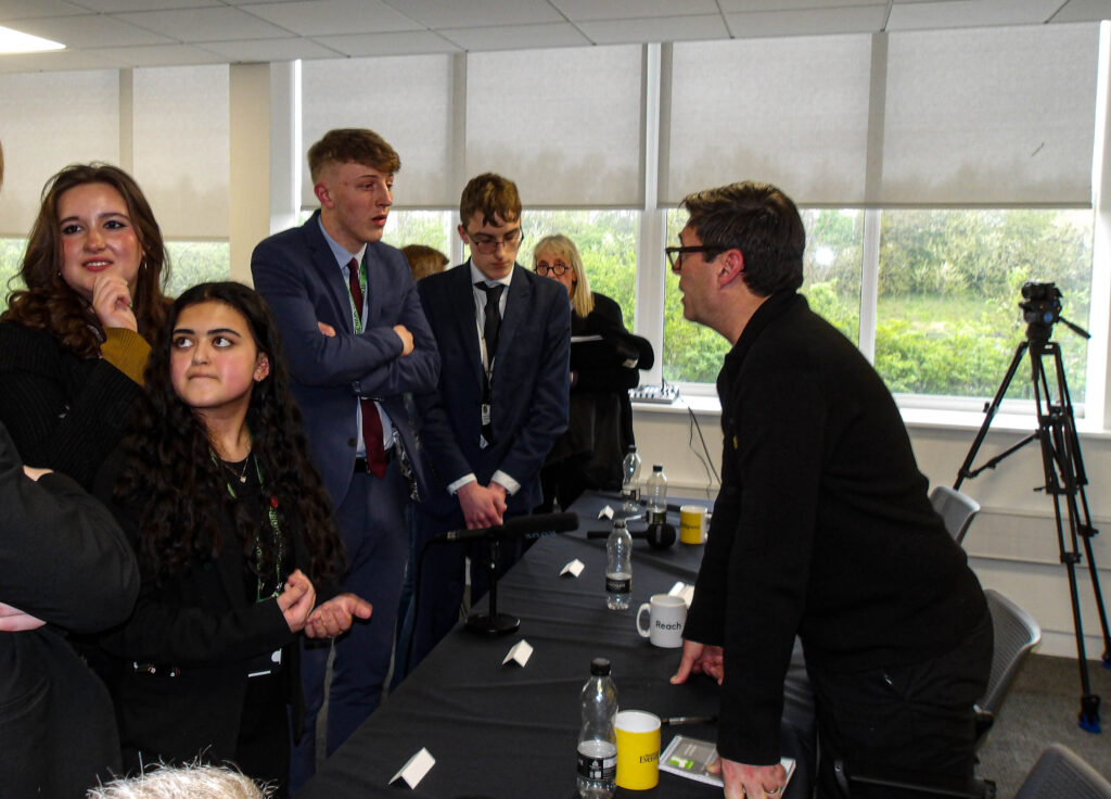 Andy Burnham leans over the hustings desk to speak to a crowd of sixth-form students in suits, who are clearly engaged with the conversation.
