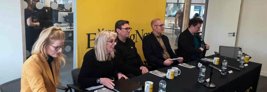 Mayoral candidates sitting at a table in the Manchester Evening News offices. From left to right: Hannah Spencer, Laura Evans, Andy Burnham, Dan Barker, and Jake Austin. Each candidate has a notebook, a mug, and a bottle of water. There is a yellow backdrop with the Manchester Evening News logo.