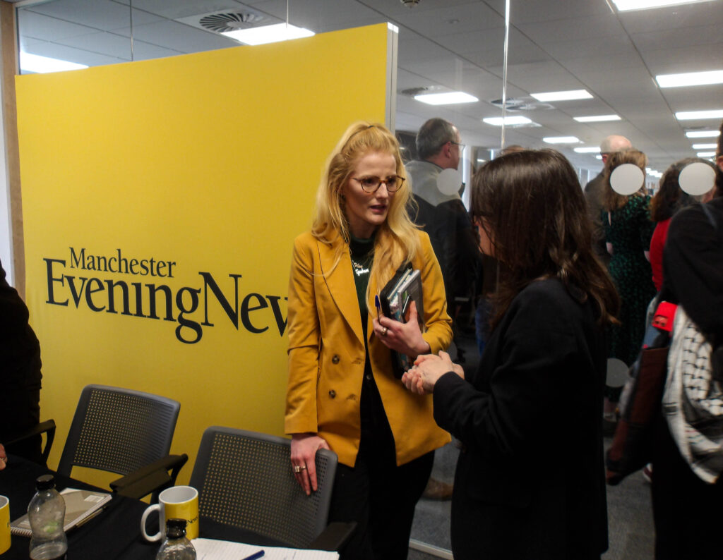 Green Party Mayoral candidate speaks to a reporter from Manchester Evening News in the Manchester Evening News offices. She is holding a pile of notebooks and engaged in conversation. Behind her is a yellow backdrop with the Manchester Evening News logo.