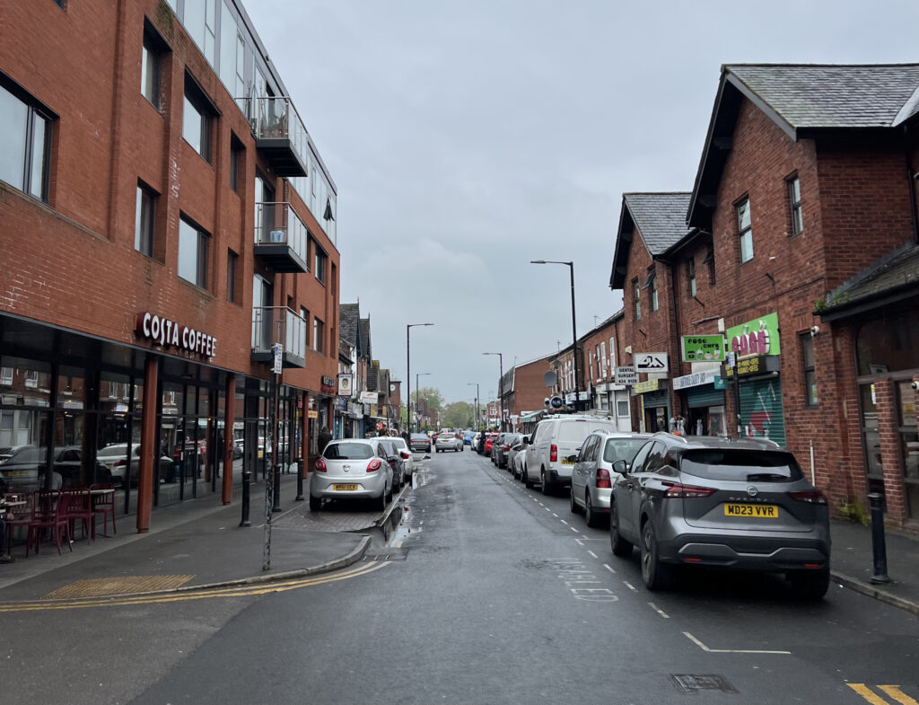 Current view of Copson Street in Withington, with cars parked at either side and businesses down the street.