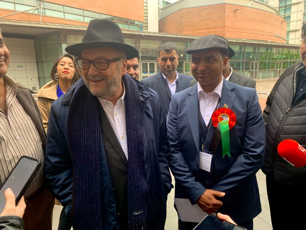 Workers' Party of Britain leader and founder George Galloway stands next to winning counsellor Shabas Sarwar. Both are smiling as Galloway talks to a reporter. They are standing outside, and the outer walls of the Manchester Conference Hall are visible. 