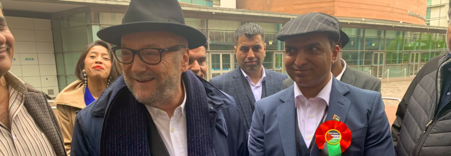 Workers' Party for Britain founder and leader George Galloway stands to the left of winning Workers' Party councillor Shabas Sarwar. Both are smiling, and Galloway is leaning towards a reporter while answering a question. They are stood outside - the walls of the Manchester Conference Hall are visible in the background.