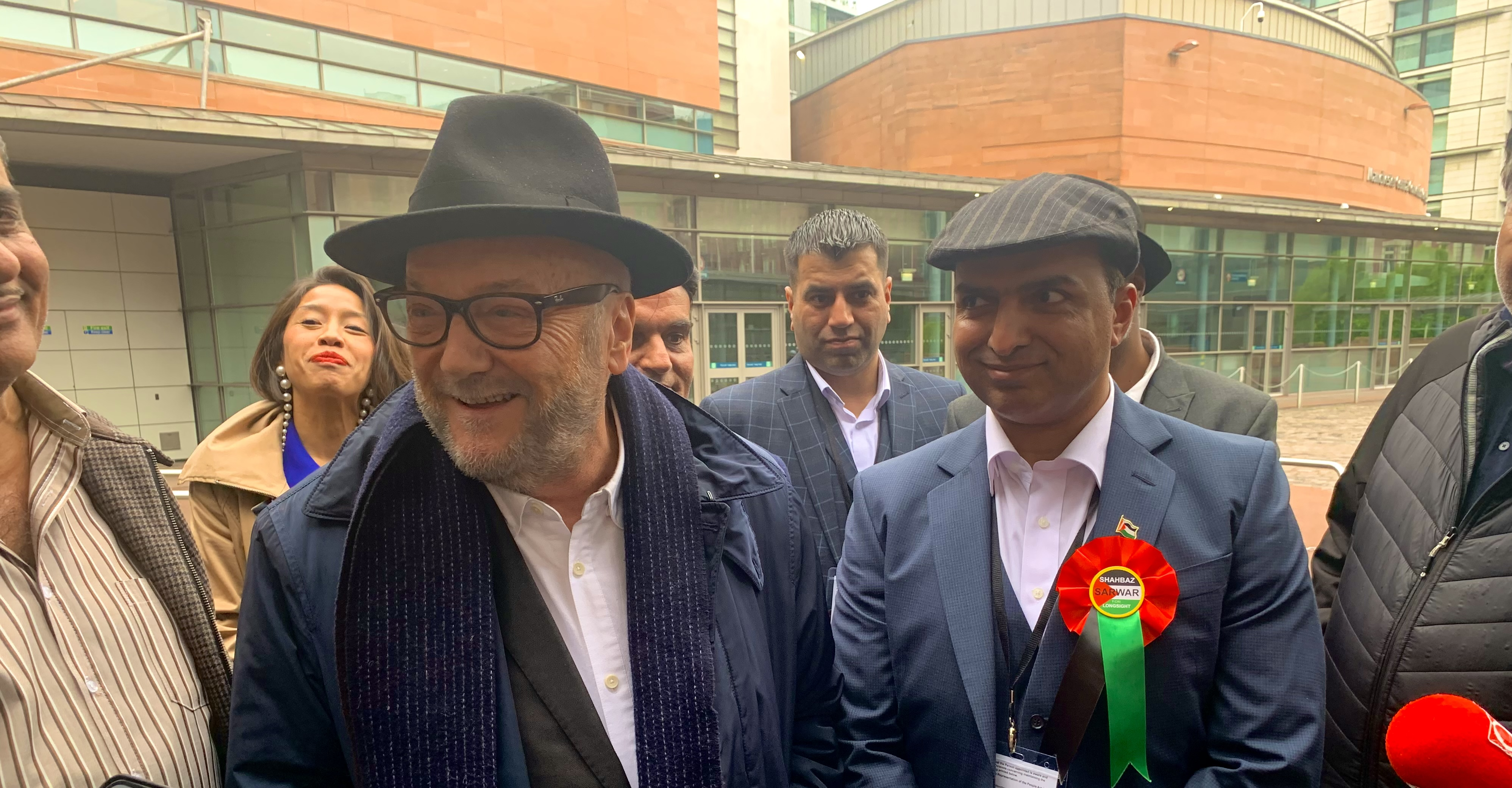 Workers' Party for Britain founder and leader George Galloway stands to the left of winning Workers' Party councillor Shabas Sarwar. Both are smiling, and Galloway is leaning towards a reporter while answering a question. They are stood outside - the walls of the Manchester Conference Hall are visible in the background.