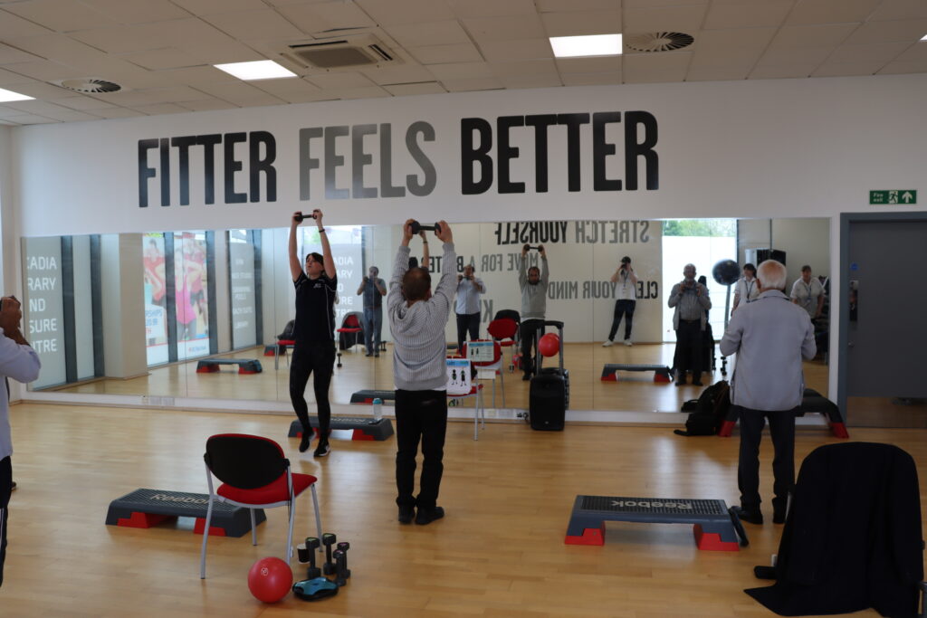 A group of people in a fitness studio holding a dumbbell above their heads.