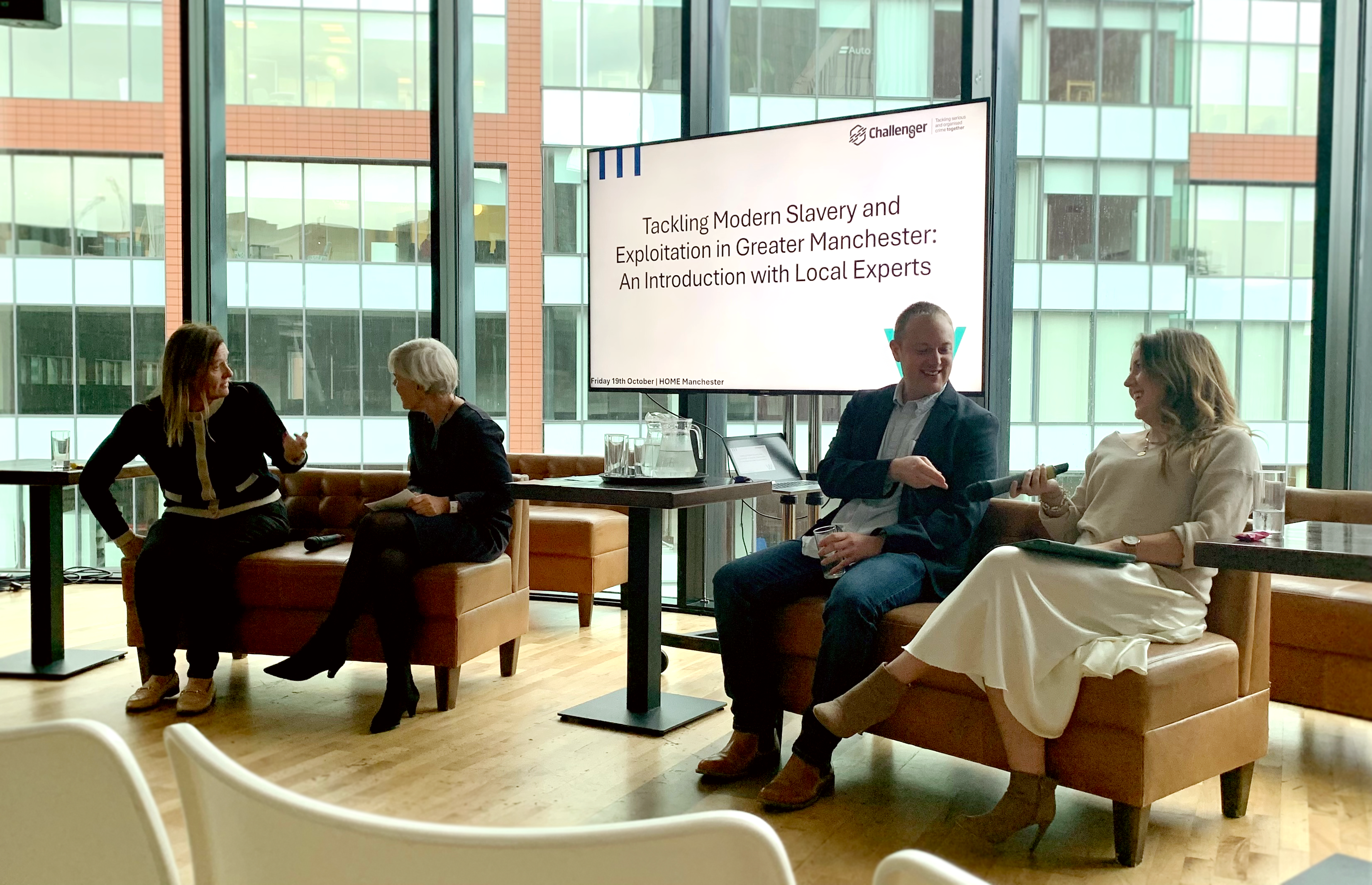 Three women and a man sit on two brown sofas in front of a digital whiteboard that reads: "Tackling Modern Slavery and Exploitation in Greater Manchester: An Introduction with Local Experts." They are sat in an open area with a glass wall behind them that faces the wall of another building. All four people are in conversation, and are well-dressed in business attire. There is a microphone on the left sofa, and the woman on the far-right of the image holds the other. In order, they are: local business lmanager Angela Hughes; Deputy Mayor of Manchester Kate Green; Dr Jon Davies from the University of Manchester; and Amy Bond from anti-slavery charity Causeway.