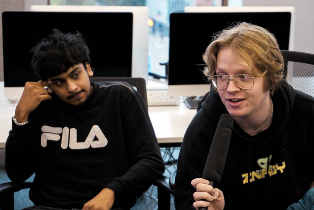 Two boys sit on the left and right side of the image, both wearing black hoodies. The boy on the left has brown skin and facial hair, and is sitting in a chair with one hand resting on his temple. The boy next to him has white skin and long, blond hair and glasses, and is speaking into a large microphone while looking off-screen.