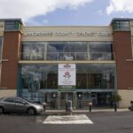 Lancashire County Cricket Club. A large building with two brick pillars, and a glass wall between with the main entrance. Above the doors is a sign with a red rose that says 'Welcome to Old Trafford Home, Home of the Lancashire County Cricket Club.'