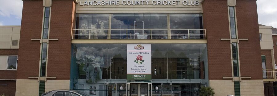 Lancashire County Cricket Club. A large building with two brick pillars, and a glass wall between with the main entrance. Above the doors is a sign with a red rose that says 'Welcome to Old Trafford Home, Home of the Lancashire County Cricket Club.'