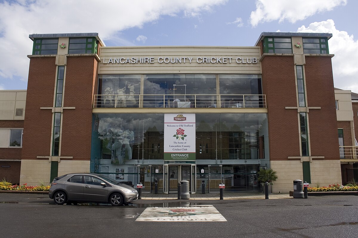 Lancashire County Cricket Club. A large building with two brick pillars, and a glass wall between with the main entrance. Above the doors is a sign with a red rose that says 'Welcome to Old Trafford Home, Home of the Lancashire County Cricket Club.'