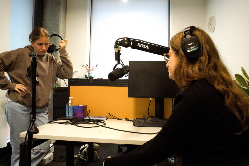 Two white women are in a brightly-lit office. The girl on the left is wearing a khaki hoodie and light blue jeans, and is standing with one hand on her hip and the other holding a pair of headphones to her ear. She is focussing intently. Sitting down at the desk on the right side of the image is a woman with ginger hair, a black jumper, and a pair of black headphones. she is leaning away from the camera and her face is not visible. There are two microphones in front of them. 