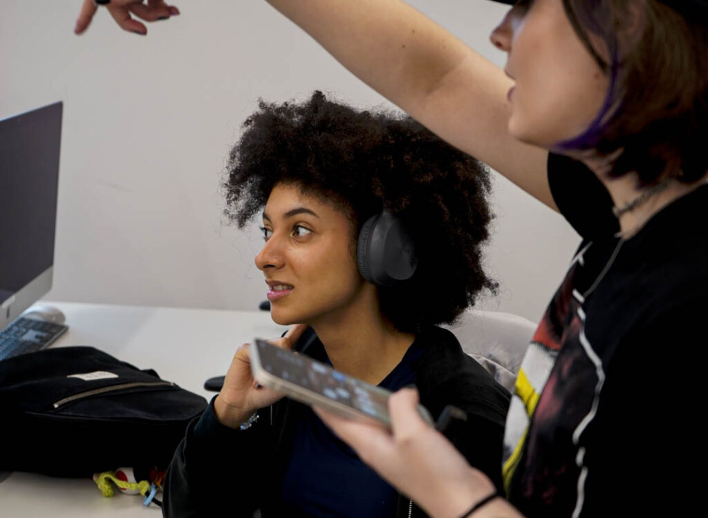 A black girl with an afro and headphones is staring intently to left at something off-screen. Her headphones are black and have a microphone attached. On the far right of the image is a white gril holding a phone in one hand and pointing to the left with another. She is speaking, and wears a black t-shirt.