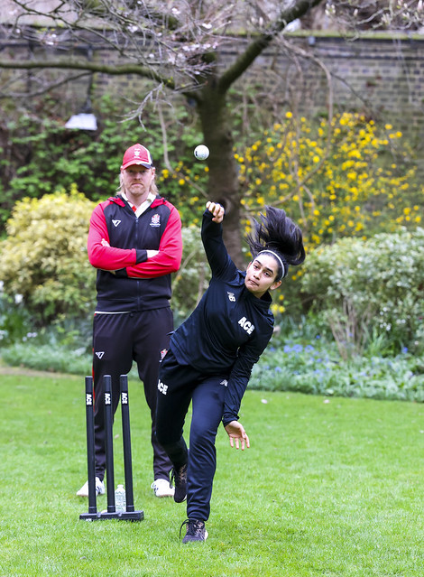 A white man and a young brown girl are in the middle of the picture, standing on grass with trees, foliage, and a brick wall in the background. The man is standing with his arms folded and facing directly into the camera, wearing a black cricket jersey with red sleeves and black trousers. there is a black cricket wick in front of him. Next to the wick, the girl stands on one leg throwing a white cricket ball. Her hair is tied back into a ponytail and she wears a black cricket jumpsuit.