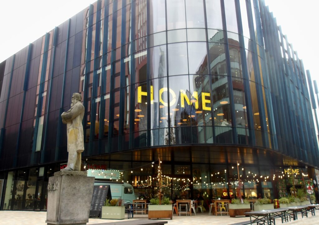 HOME Gallery, Manchester. A tall building of tinted glass, square-shaped with curved edges. There are tables and chairs outside the entrance lit by strings of lights overhead. To the left is a stone statue of a man in a 19th-century suit with his arms folded, stood on a plinth. The statue is facing left in profile.