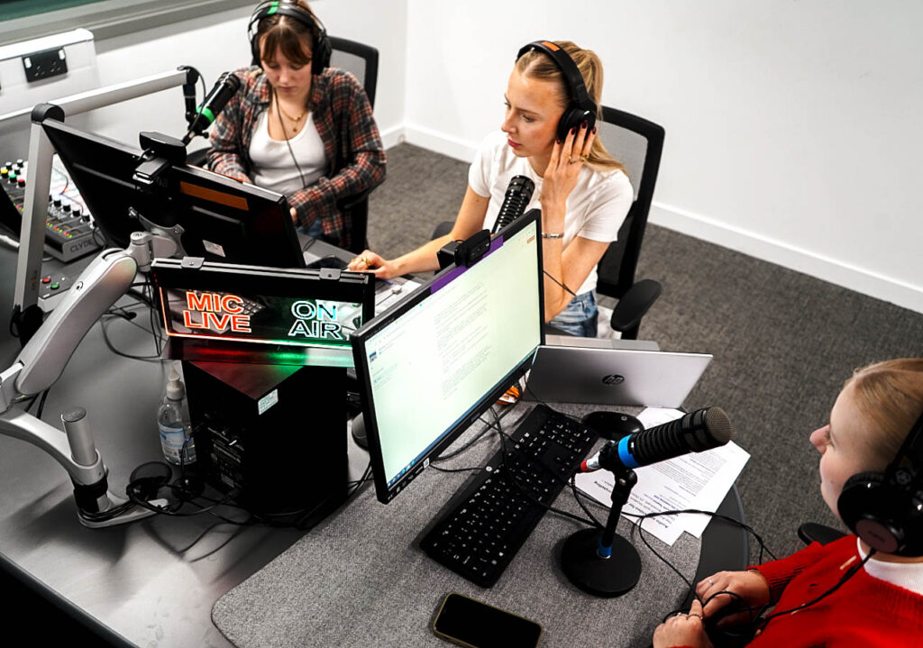 Three girls sat around a table in the bottom left corner, each with a computer and a microphone. In the middle of the table is a red and green sign which says LIVE ON AIR. The girl in the upper right corner is white and blonde, with a fringe, a white vest, and a checked shirt, and is wearing headphones while typing. The girl in the middle is white, with blonde hair in a ponytail and a white top. She is holding her headphones to her ear with one hand while she types. The girl in the bottom right corner is white, with blonde hair and headphones, and is wearing a bright red jumper. She is reading a script on the computer, with the words out of focus in the image.