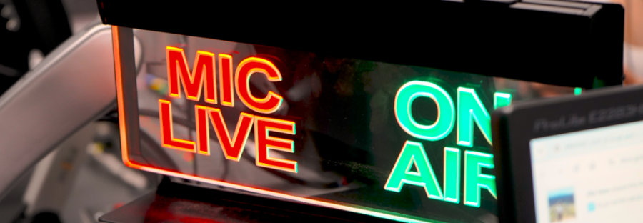 An ON AIR sign in a radio studio. The picture is extremely zoomed-in, with a sign that says MIC LIVE in glowing red on the left side, and ON AIR in glowing green on the right.