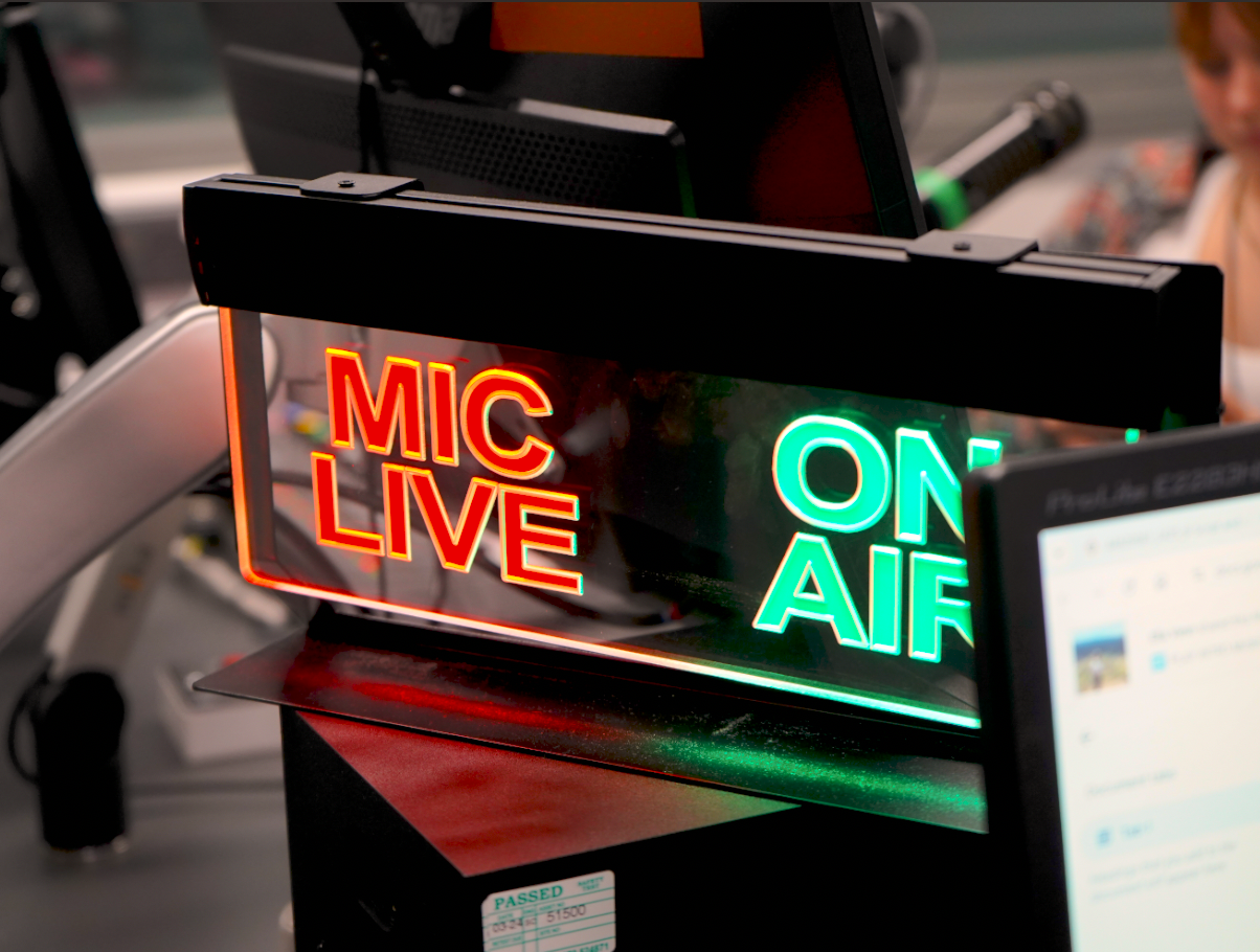 An ON AIR sign in a radio studio. The picture is extremely zoomed-in, with a sign that says MIC LIVE in glowing red on the left side, and ON AIR in glowing green on the right.