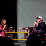 A white woman in a hot pink dress with a microphone sits on an armchair opposite Jamie Laing, a white man in a white cap who is also holding a microphone. Between them is a table full of jugs of water, and they are sat under spotlights on a stage. Jamie is talking.