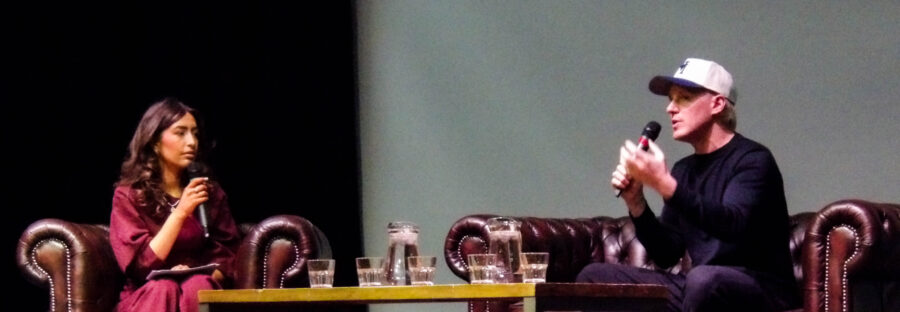 A white woman in a hot pink dress with a microphone sits on an armchair opposite Jamie Laing, a white man in a white cap who is also holding a microphone. Between them is a table full of jugs of water, and they are sat under spotlights on a stage. Jamie is talking.