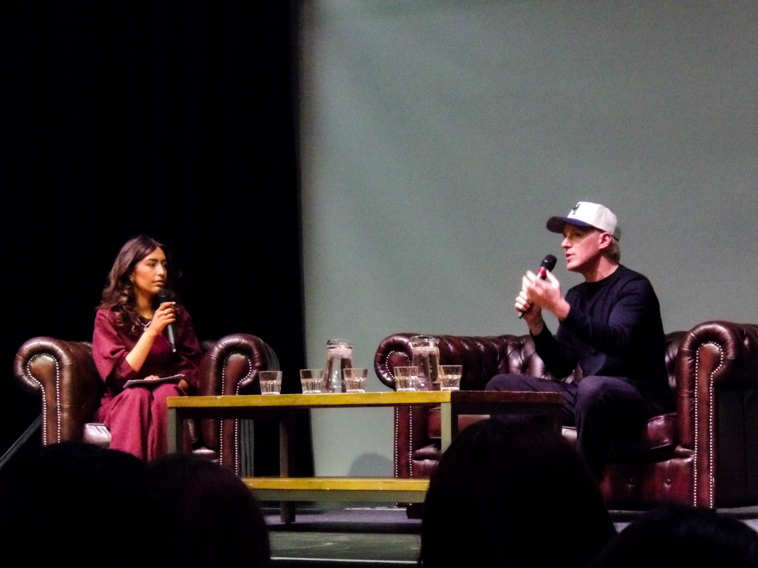 A white woman in a hot pink dress with a microphone sits on an armchair opposite Jamie Laing, a white man in a white cap who is also holding a microphone. Between them is a table full of jugs of water, and they are sat under spotlights on a stage. Jamie is talking.