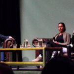 A white man in a blue jacket interviews a brown woman and white man on a sofa. All three are holding microphones, and a table filled with water jugs sits between them.