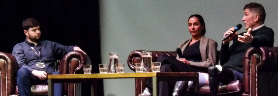 A white man in a blue jacket interviews a brown woman and white man on a sofa. All three are holding microphones, and a table filled with water jugs sits between them.