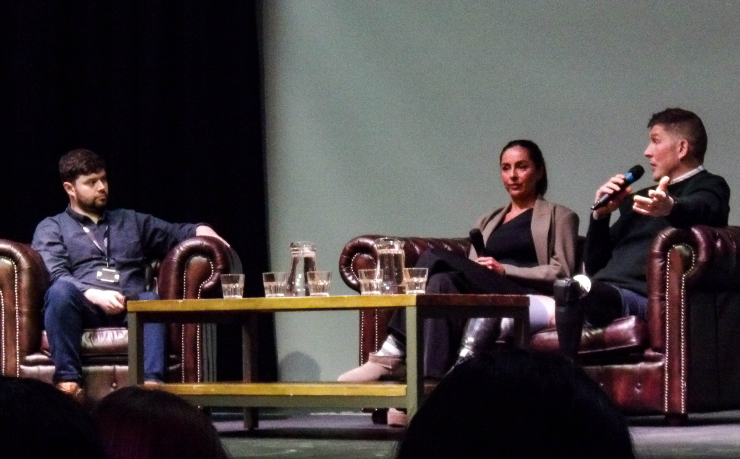 A white man in a blue jacket interviews a brown woman and white man on a sofa. All three are holding microphones, and a table filled with water jugs sits between them.