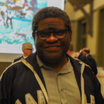 Gary Younge, a Black man wearing a grey button-up shirt and navy blue hoodie, is smiling directly into the camera. In the background is a large lecture theatre with rows of fold up chairs and a large screen with a blurred painting. There are some blurred figures in the background.