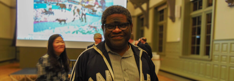 Gary Younge, a Black man wearing a grey button-up shirt and navy blue hoodie, is smiling directly into the camera. In the background is a large lecture theatre with rows of fold up chairs and a large screen with a blurred painting. There are some blurred figures in the background.