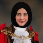 Cllr. Tafheen Sharif. A British-Kashmiri woman in a black hijab, white collar, red cloak, and gold chains. She is smiling at the camera