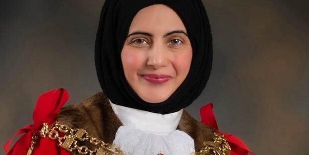 Cllr. Tafheen Sharif. A British-Kashmiri woman in a black hijab, white collar, red cloak, and gold chains. She is smiling at the camera