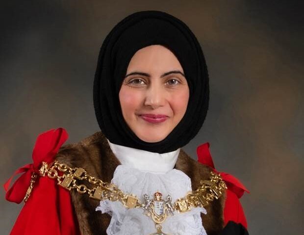 Cllr. Tafheen Sharif. A British-Kashmiri woman in a black hijab, white collar, red cloak, and gold chains. She is smiling at the camera