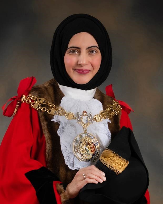 Cllr. Tafheen Sharif. A British-Kashmiri woman in a black hijab, white collar, red cloak, and gold chains. She is smiling at the camera