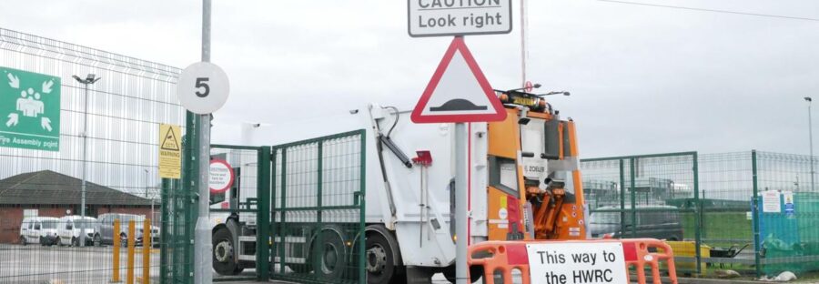 Exterior gates of Bolton recycling facility. A bin lorry is moving forward past a sign that reads "This way to the HWRC"