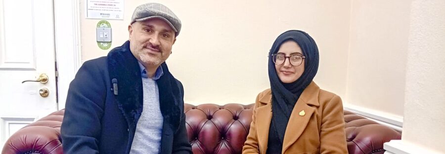 Faraz Aryan, a man in a light blue shirt, dark blue jacket, and flat cap, sits on a red leather sofa next to Cllr. Tafheen Sharif, a British-Kashmiri woman in a blue hijab and orange leather jacket. Both are smiling into the camera.