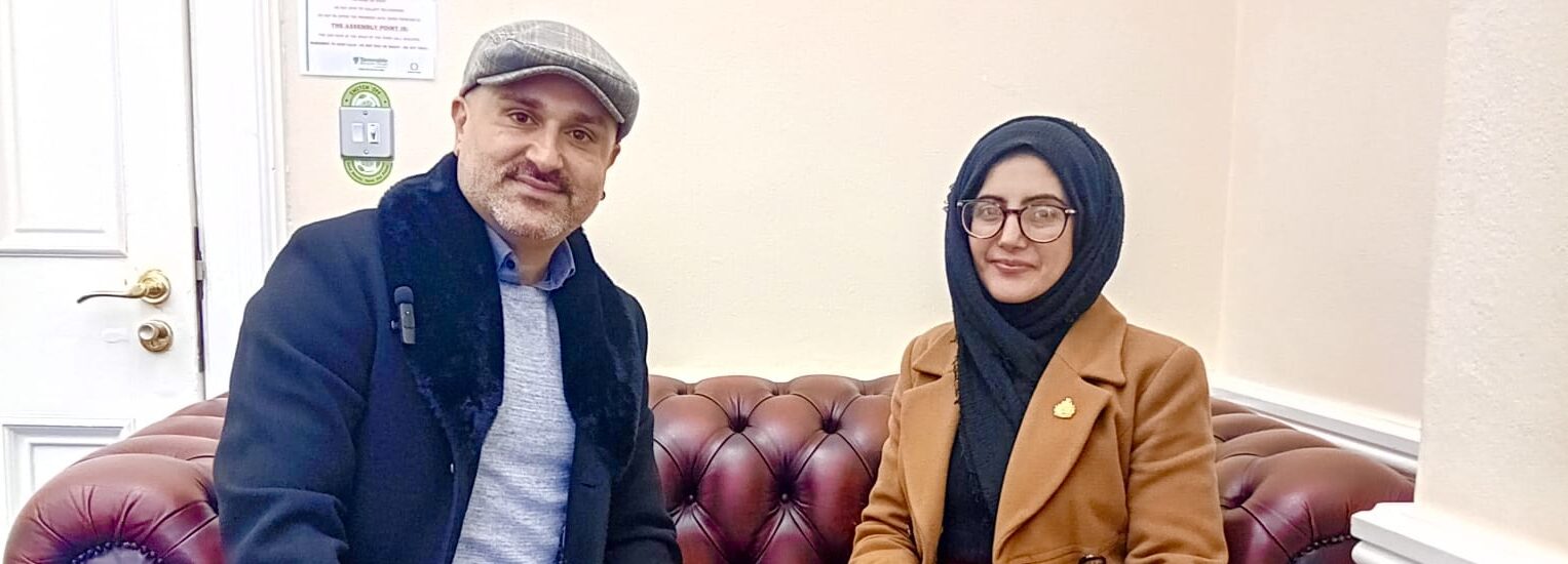 Faraz Aryan, a man in a light blue shirt, dark blue jacket, and flat cap, sits on a red leather sofa next to Cllr. Tafheen Sharif, a British-Kashmiri woman in a blue hijab and orange leather jacket. Both are smiling into the camera.