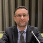 Dr Haytham Alhamwi is sitting with his arms folded on a table. He has a screen and microphone in front of him, and a sign that says Haytham Alhamwi in front of him. He is looking into the camera and smiling.