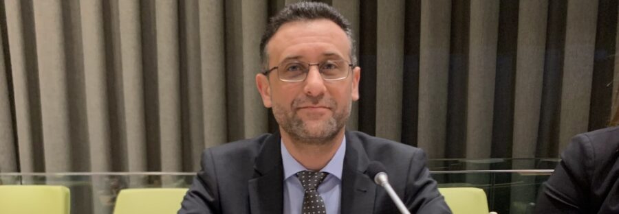 Dr Haytham Alhamwi is sitting with his arms folded on a table. He has a screen and microphone in front of him, and a sign that says Haytham Alhamwi in front of him. He is looking into the camera and smiling.